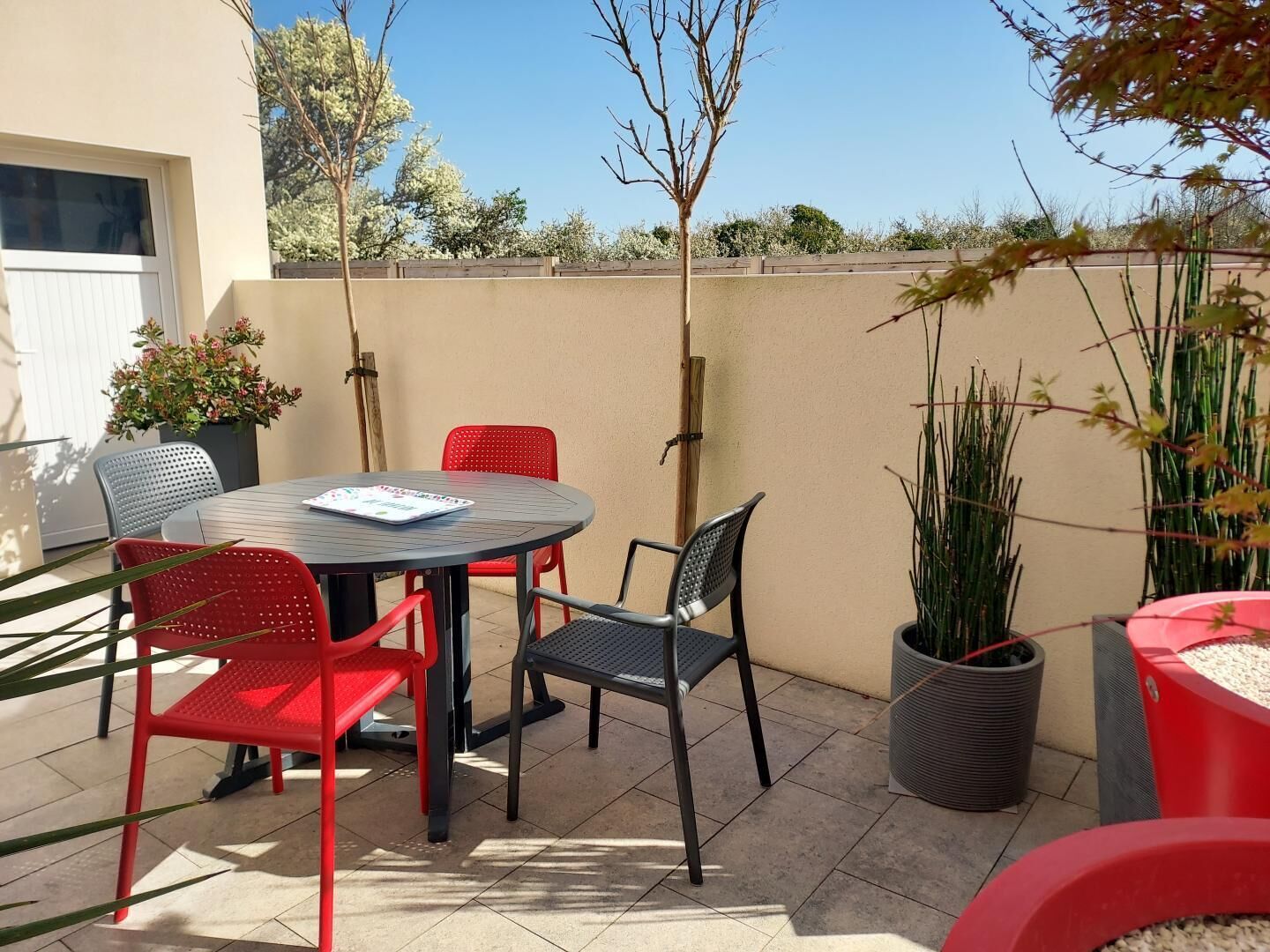 En terrasse med et rundt bord og fire stole, omgivet af plantekasser og en beige væg.