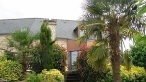Une maison avec une façade en pierre, un chemin d’accès en pierre et une végétation luxuriante comprenant des palmiers et des arbustes.
