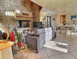 A living room with a stone fireplace, a television, a sofa, and a dining area with a table and chairs. - Le Gîte du Procureur (MONTERBLANC)