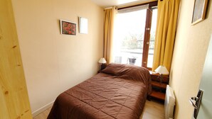 Una habitación pequeña con cama individual, suelo de madera, ventana con cortinas amarillas, radiador y dos lámparas de pared.