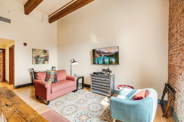A living room with a wooden ceiling, a pink sofa, a blue armchair, a patterned rug, and a painting of a mountain landscape.