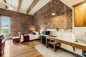 A compact kitchen with a brick wall, a bed with a red headboard, and a wooden ceiling.