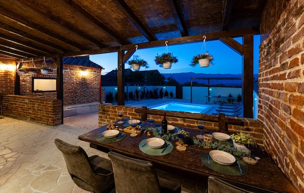 An outdoor dining area with a brick wall, wooden beams, hanging planters, and a view of a swimming pool and mountains.