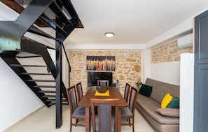 A dining area with a wooden table and chairs, a stone wall with a colorful painting, a sofa with cushions, and a black spiral staircase.
