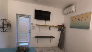Una habitación pequeña y funcional con una cama, un televisor montado, un equipo de aire acondicionado, un cuadro y un estante con objetos personales.