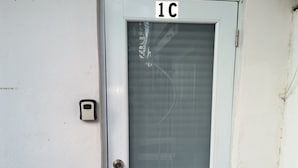 Una puerta con un panel de vidrio esmerilado, una placa con el número “1C” y un pequeño intercomunicador a un lado.