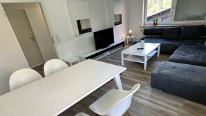 Un salon moderne avec une table à manger blanche, des chaises blanches, un canapé gris et un téléviseur fixé au mur.