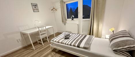 Una habitación pequeña y limpia con una cama, un escritorio y una silla. Tiene una ventana con cortinas, un radiador y una lámpara fijada a la pared.