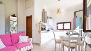 Un salon lumineux avec un canapé rose, un coin repas avec des chaises blanches et une cuisine équipée d’appareils modernes.