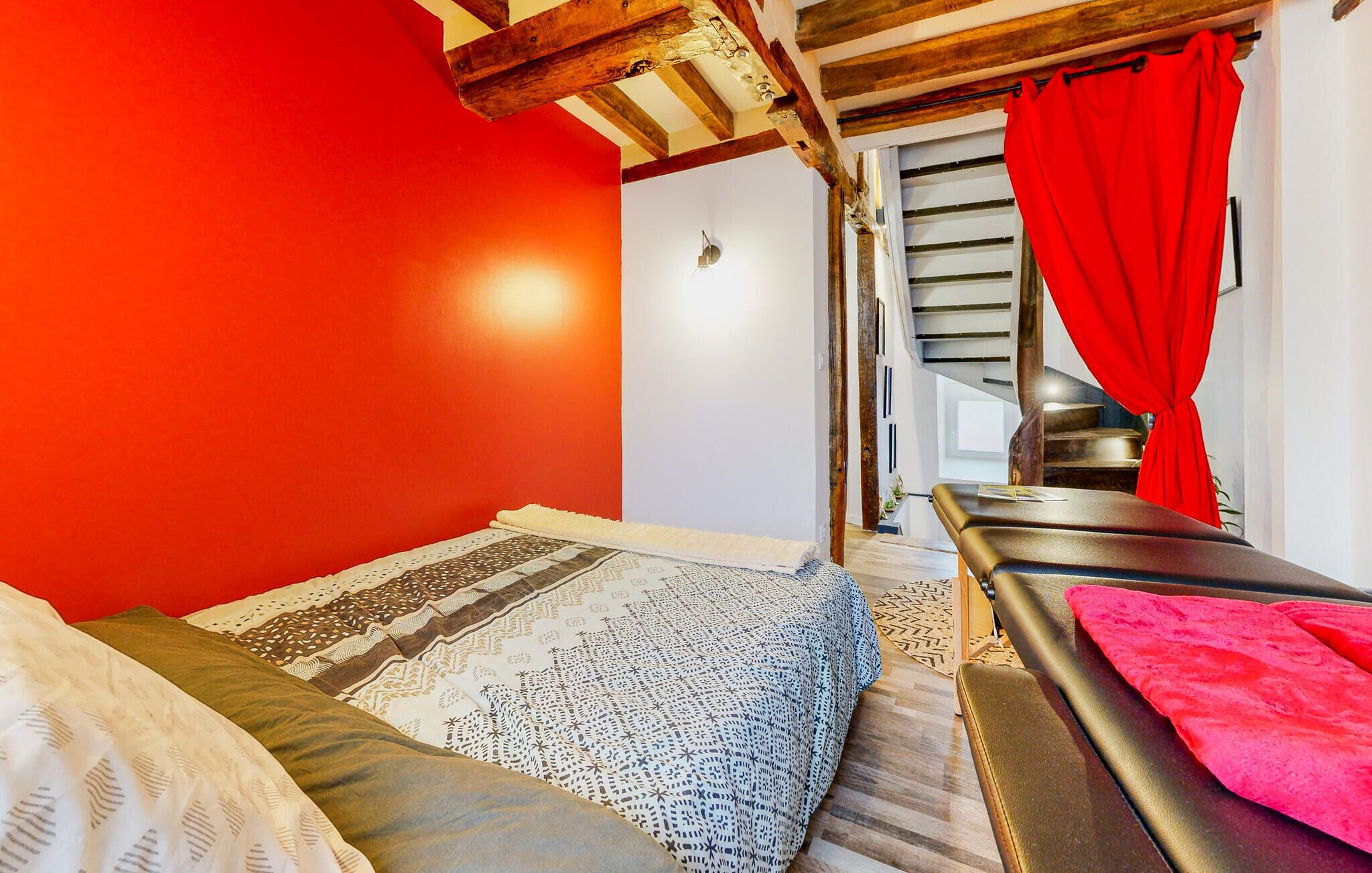 A room with a red wall, a bed with patterned bedding, a massage table with a red towel, and a staircase with a red curtain.