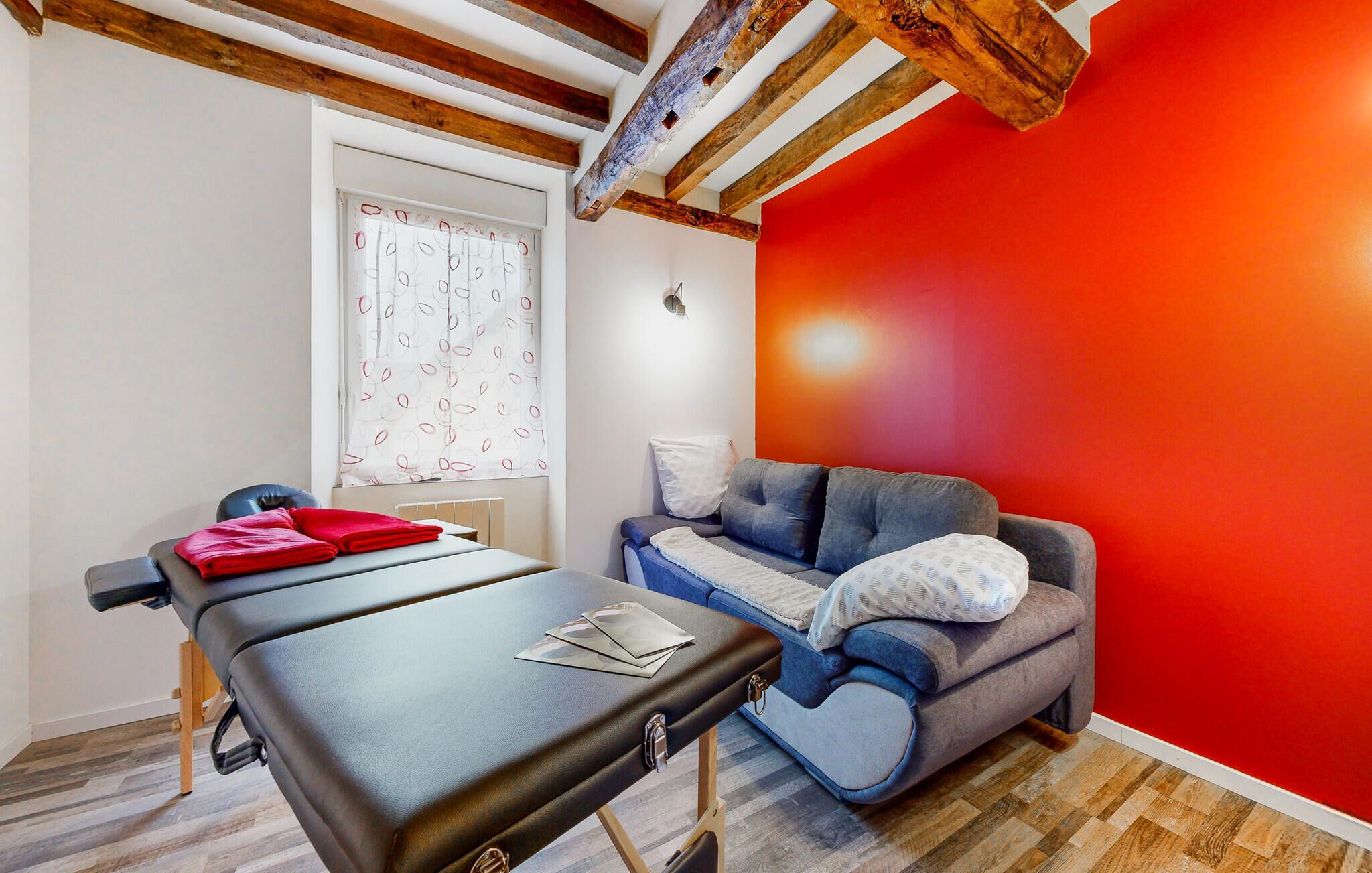 Een kamer met een massagetafel, een grijze bank en een rode accentmuur.