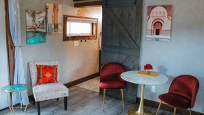 Une pièce avec une porte en bois, une petite table ronde surmontée d’un vase, deux chaises rouges, un fauteuil à motifs et une petite table recouverte d’une nappe à motifs.