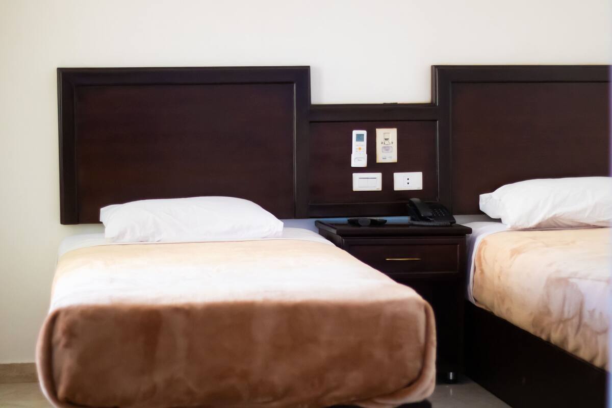 A hotel room with two single beds, a nightstand with a phone, and a wall-mounted control panel.