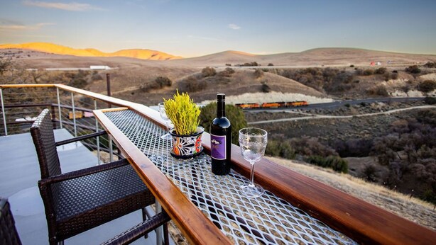 Una mesa con una botella de vino, una copa y una planta en maceta en un balcón con vistas a un paisaje montañoso.