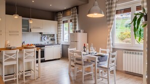 A modern kitchen with white cabinetry, a dining area with white chairs, and a view of greenery outside. - Graziella by Interhome (Leggiuno)