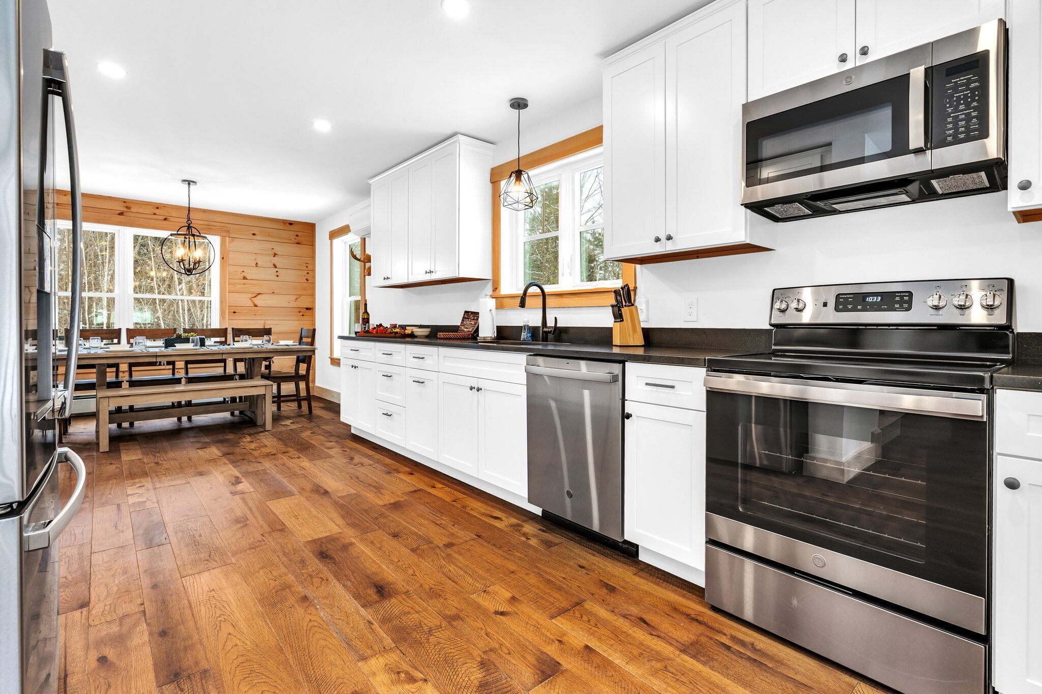 A modern kitchen with white cabinets, stainless steel appliances, a wooden floor, and a dining area with a bench.
