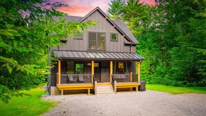 A two-story wooden cabin with a porch, surrounded by lush greenery. - NEW Chalet 6 minutes from Sunday River! Hot tub, Sauna, Sleeps 12, Dog Friendly! (Bethel)