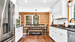 A modern kitchen with white cabinets, stainless steel appliances, a wooden dining table, and a large window. - NEW Chalet 6 minutes from Sunday River! Hot tub, Sauna, Sleeps 12, Dog Friendly! (Bethel)