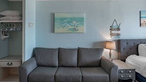 A bedroom with a grey sofa, a bedside table with a lamp, a bed, a painting of a beach, and a wall-mounted sign. - Coastal Retreat with Ocean Views | Driftwood 1608 (Myrtle Beach)