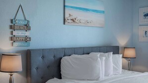 A bedroom with a bed, two bedside lamps, a wall-mounted artwork of a beach scene, and a series of wooden signs with beach-related text. - Coastal Retreat with Ocean Views | Driftwood 1608 (Myrtle Beach)