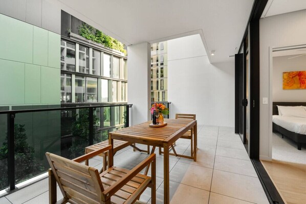 Ein Balkon mit Holztisch und -stühlen, Blick ins Grüne und ein modernes Interieur mit Bett und Kunstwerken.