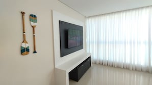 A modern living room with a flat-screen TV mounted on a white console, two decorative paddles on the wall, and a large window with sheer curtains. - Refúgio Praiano em Bombinhas (Centro Bombinhas)