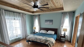 Un dormitorio con techo de madera, una cama con colcha azul y beige, dos mesitas de noche con lámparas y una ventana con cortinas.