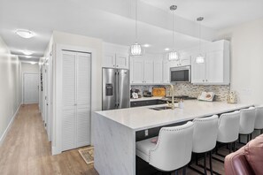 A modern kitchen with white cabinetry, stainless steel appliances, pendant lighting, and a bar area with stools. - Located in Casa Del Mila Oro, ✨1st‑Floor Condo • 2 bedroom • Steps to the Pool ✨ (Osoyoos)