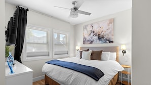 A hotel room with a bed, a ceiling fan, two windows with blinds, a wall-mounted abstract painting, bedside lamps, and a small wooden side table. - Located in Casa Del Mila Oro, ✨1st‑Floor Condo • 2 bedroom • Steps to the Pool ✨ (Osoyoos)