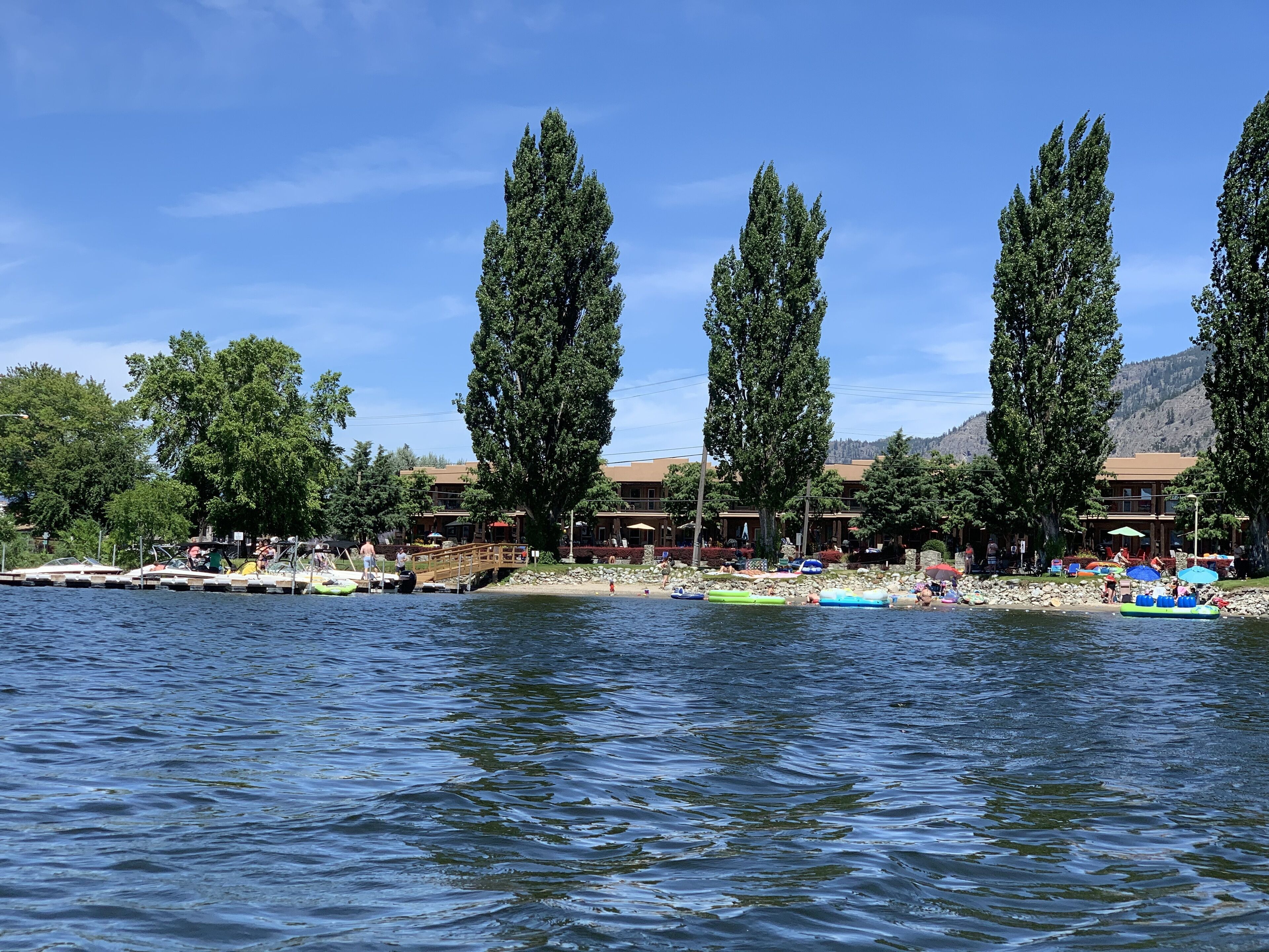A lakeside resort with docked boats, a wooden pier, and a row of tall trees.