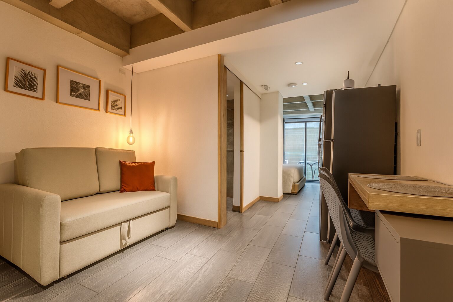 A modern living room with a sofa, a coffee table, and a refrigerator. There are framed pictures on the wall and a window with a view.