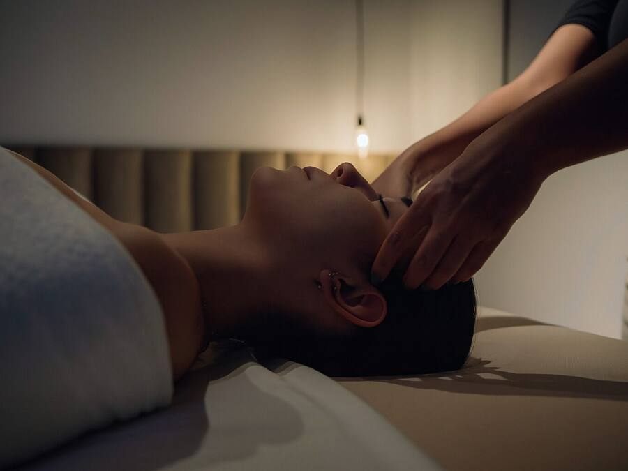Una persona recostada en una cama con una diadema, otra persona atendiéndola, y una lámpara de noche al fondo.