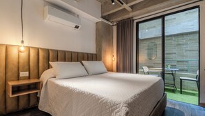Un dormitorio moderno con una cama grande, una mesita de noche, un banco empotrado, un aire acondicionado de pared y un ventanal corredero que da a un espacio exterior con mesa y sillas.