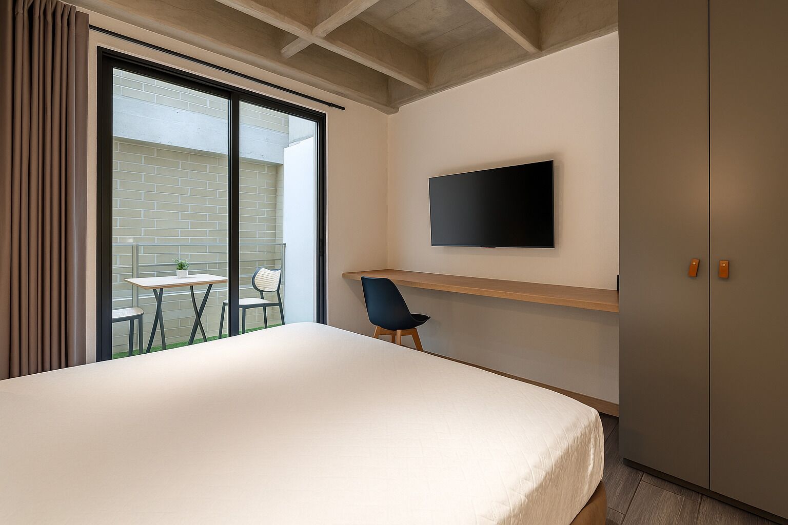 Habitació d'hotel amb un llit gran, un televisor muntat a la paret, una taula petita amb cadires i un balcó amb vista a un edifici de maó.
