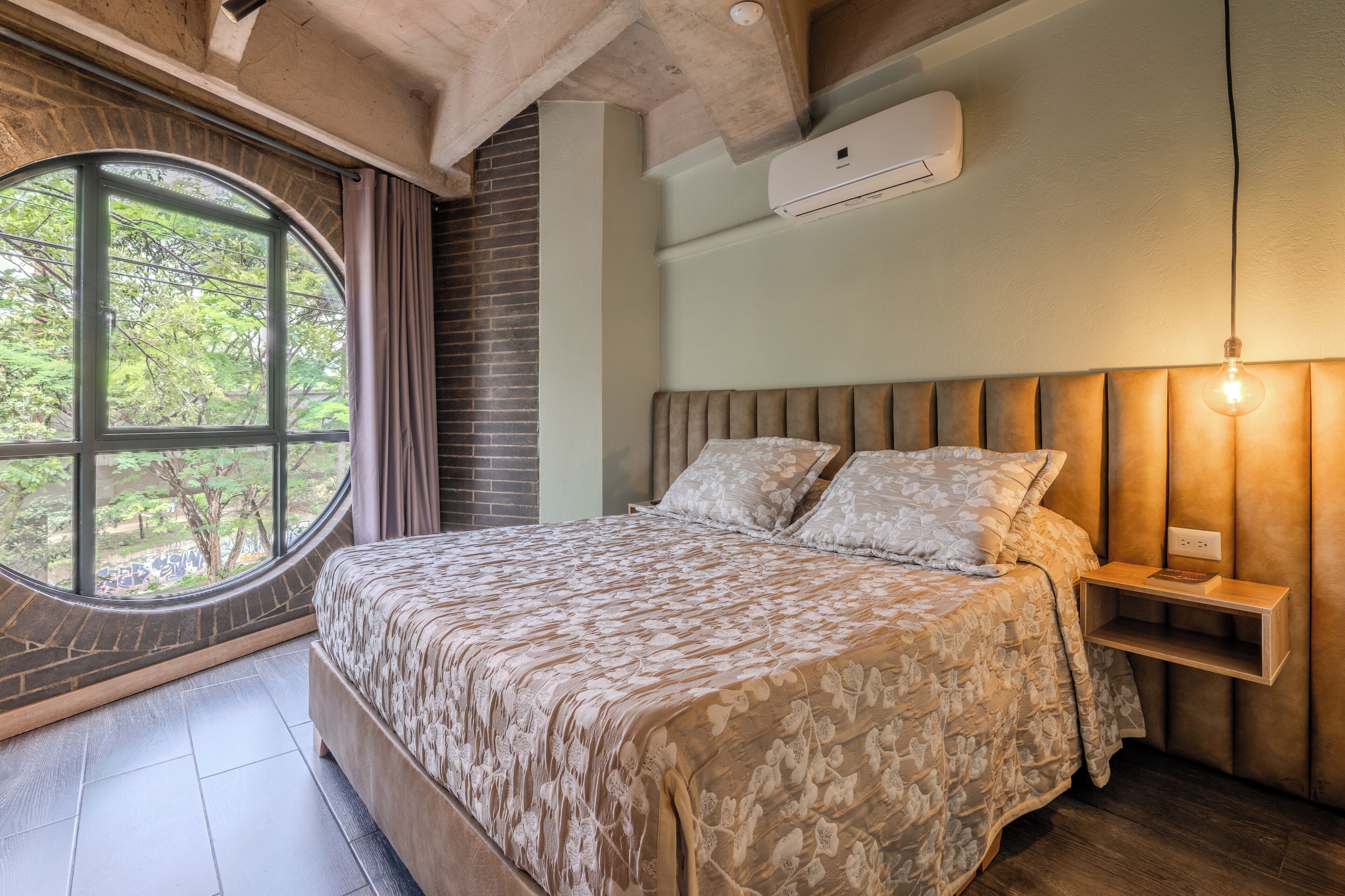 Un dormitorio con un ventanal grande, una cama con cubrecama estampado, una mesita de noche con lámpara y un aire acondicionado instalado en la pared.