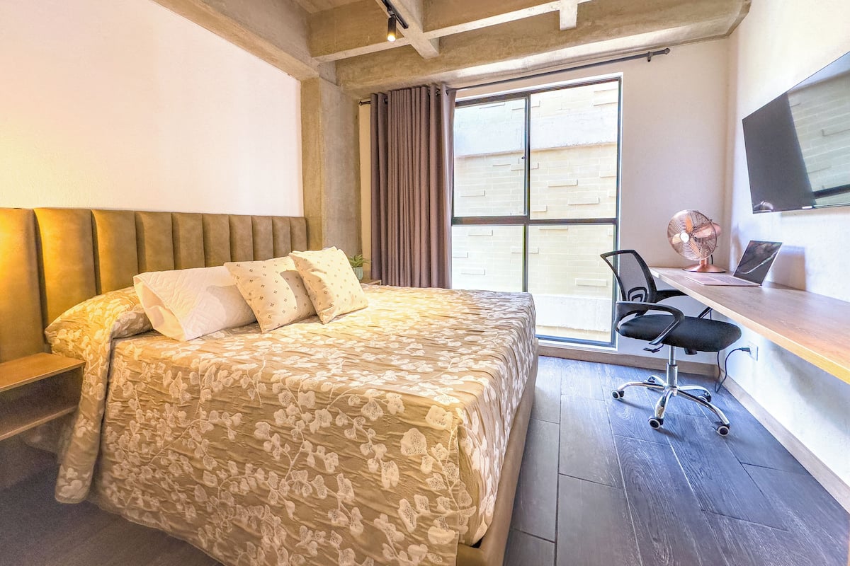 Une chambre à coucher avec un lit, un bureau avec un ordinateur portable, une chaise et une grande fenêtre.