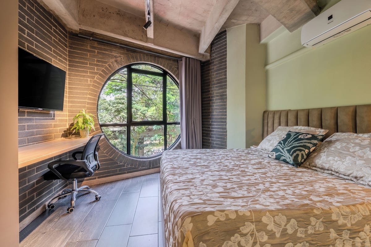 Une chambre avec un grand lit, un bureau avec une chaise, un téléviseur fixé au mur et une fenêtre avec des rideaux.