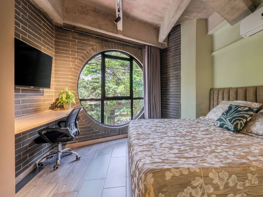 A bedroom with a large bed, a desk with a chair, a TV mounted on the wall, and a window with curtains.
