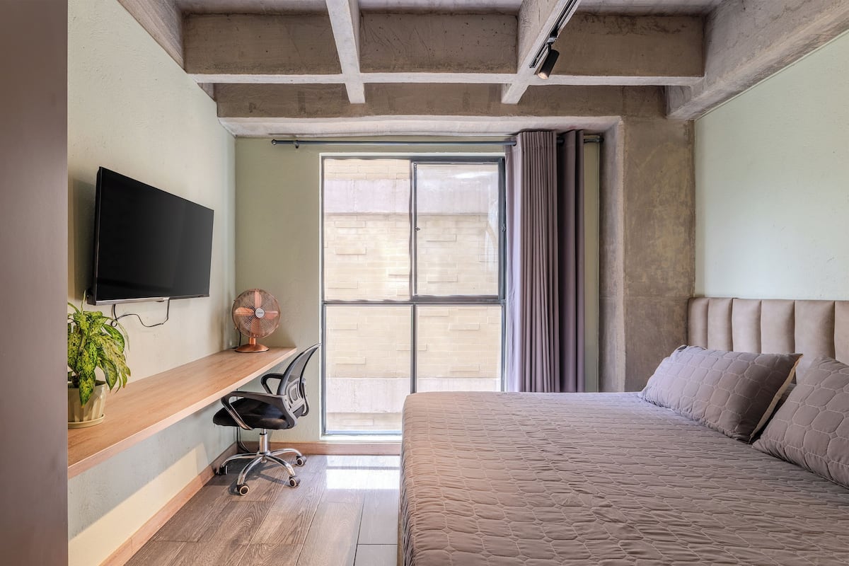 Une chambre à coucher avec un lit, un bureau avec une chaise, une télévision et une fenêtre avec des rideaux.