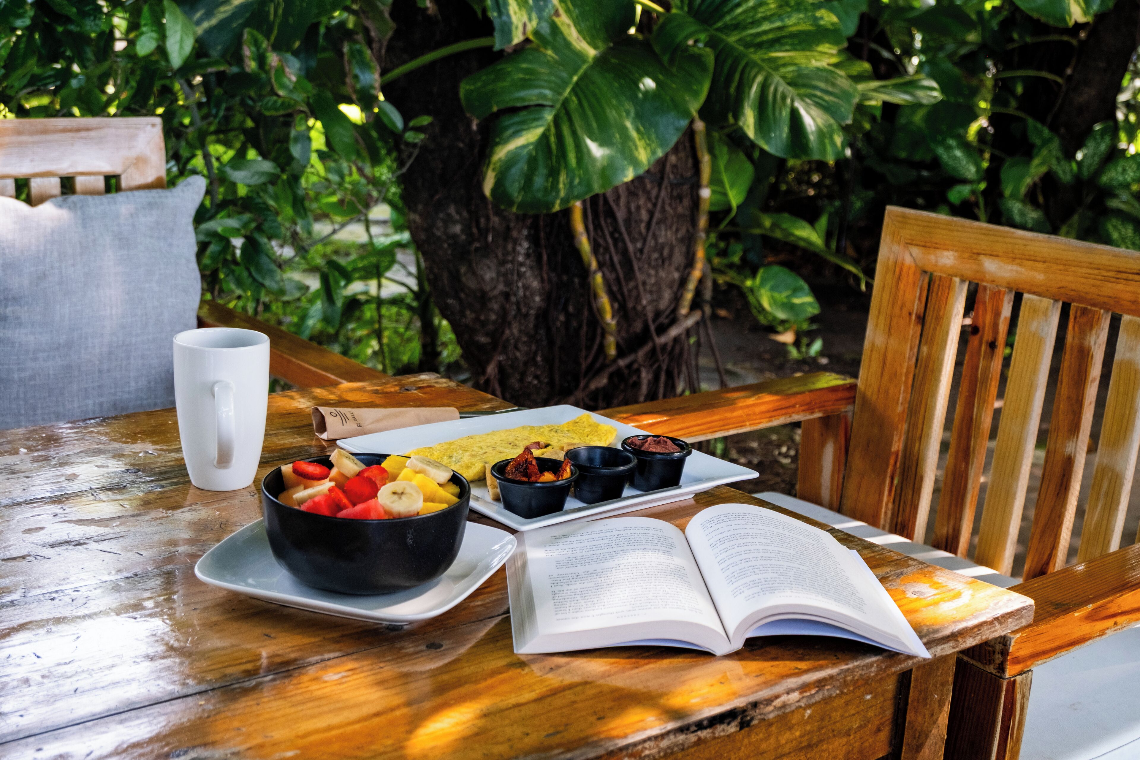 A wooden table with a bowl of fruit, a plate of food, a white mug, and an open book.