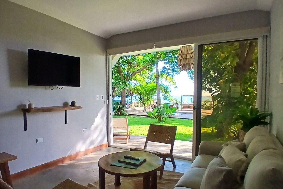Una sala de estar con un sofá, una mesa de centro, un televisor montado en la pared y una puerta corrediza de vidrio que da a un espacio exterior con césped y sillas.