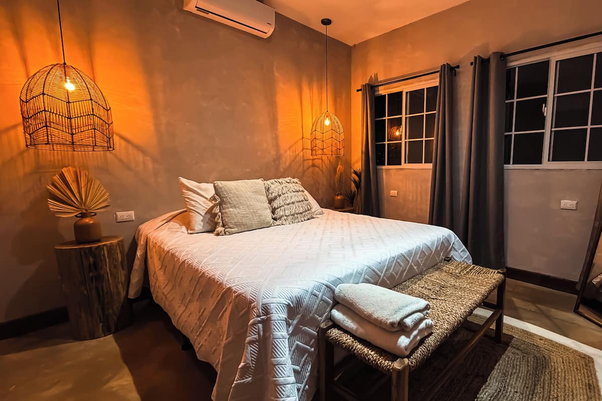 Un dormitorio con cama, lámparas de noche, una ventana con cortinas, un espejo y una pequeña mesa con un florero.