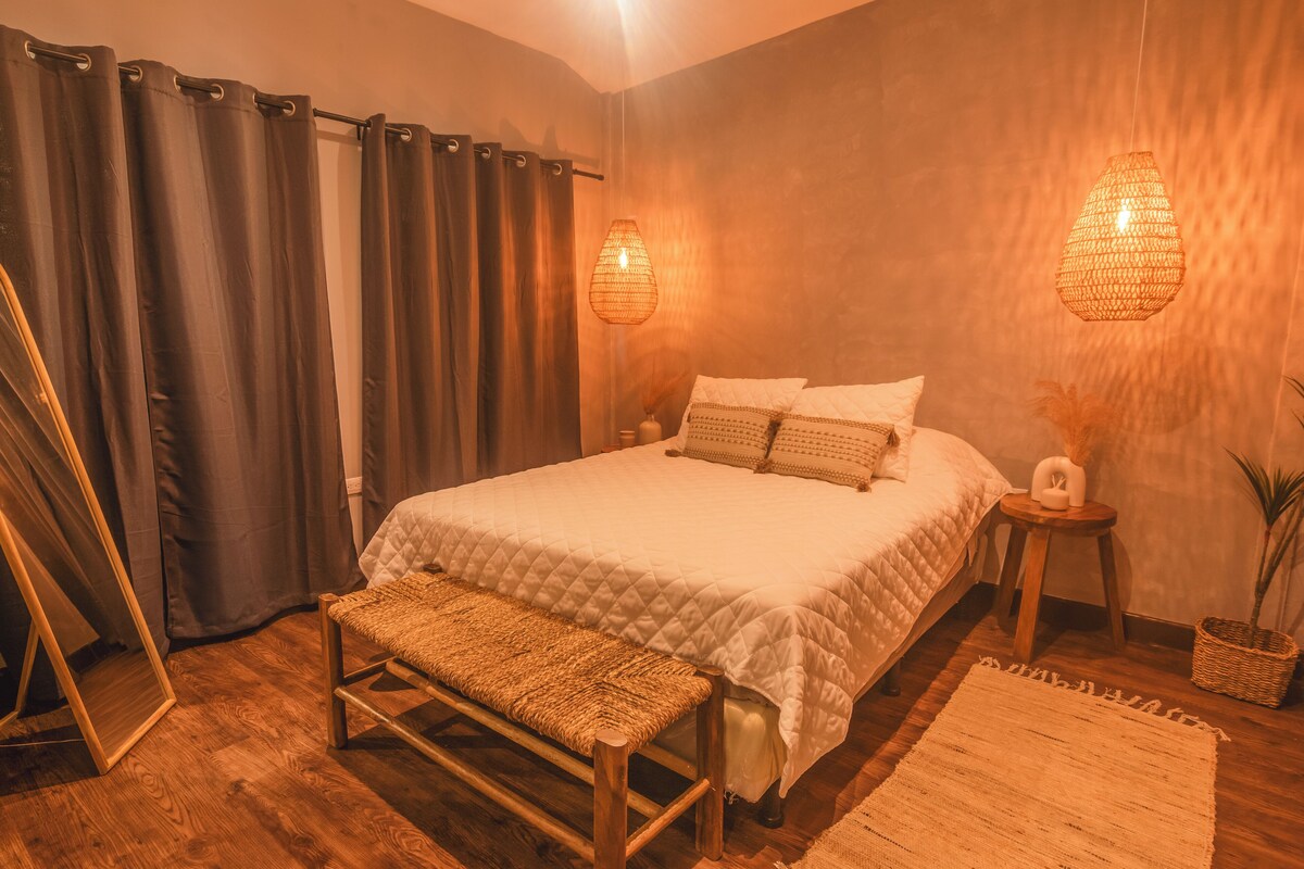 Un dormitorio con cama, banca, mesita de noche, espejo, cortinas y una lámpara colgante.
