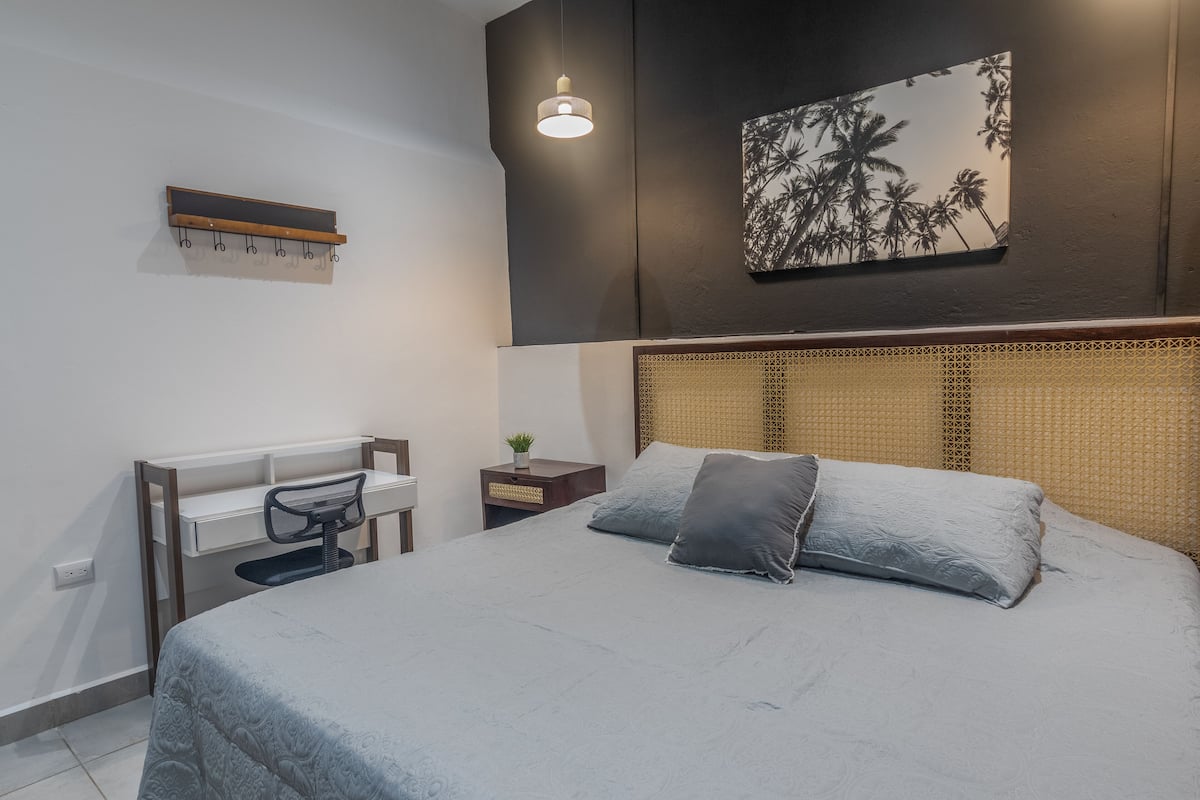 Un dormitorio con cama, escritorio, silla, mesita de noche, estante en la pared y una obra de arte.