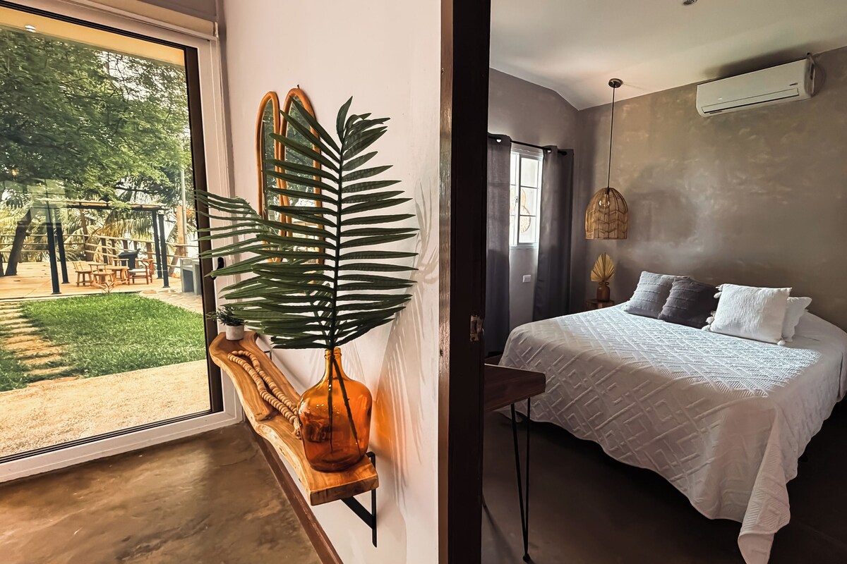 Un dormitorio con una cama grande, un banco de madera, una planta en maceta y vistas de un jardín a través de una puerta de cristal.