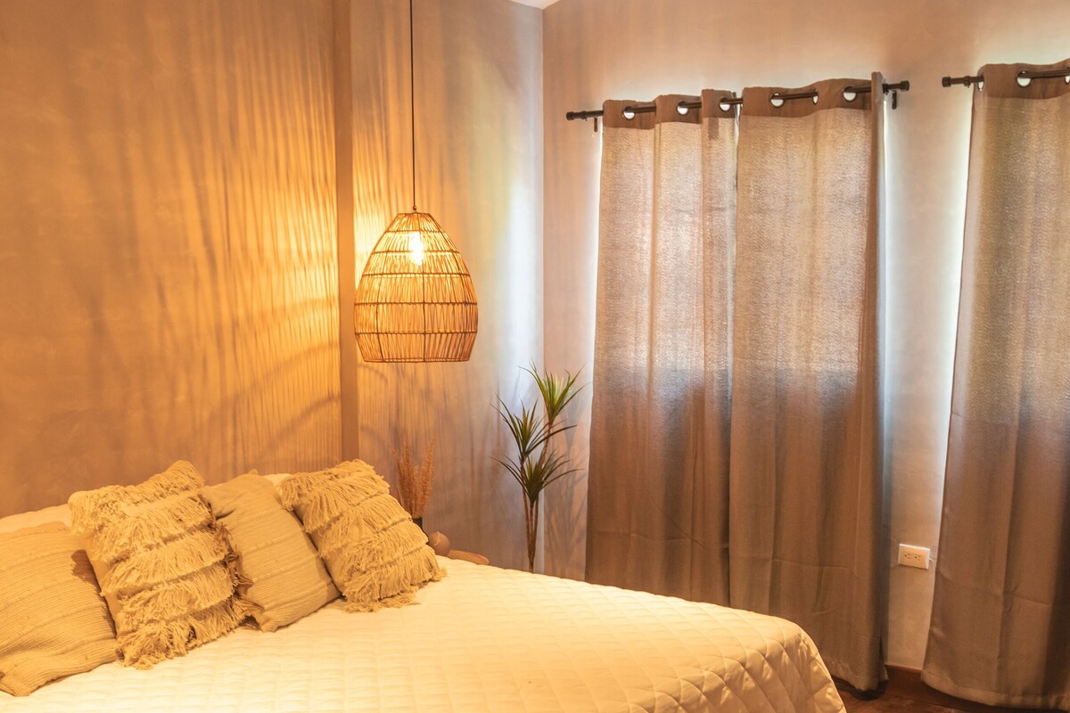 Un dormitorio con una cama grande, un banco, una lámpara colgante y cortinas.