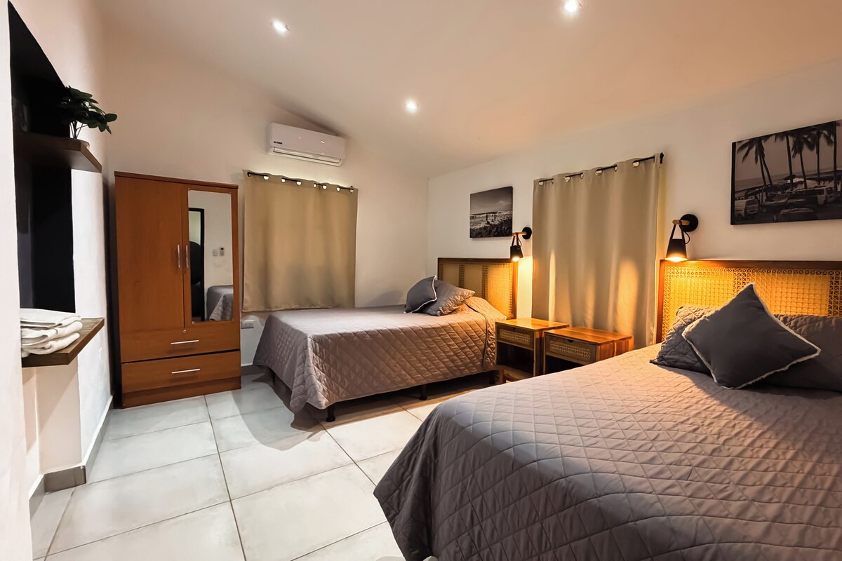 Una habitación de hotel con dos camas, un armario, un aire acondicionado, una mesita de noche con lámpara y un cuadro en la pared.