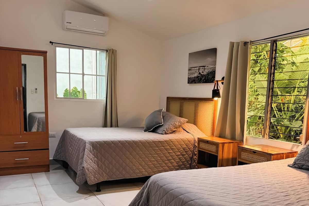 Habitación de hotel con dos camas, un armario, aire acondicionado, una ventana con cortinas y una mesita de noche con lámpara.