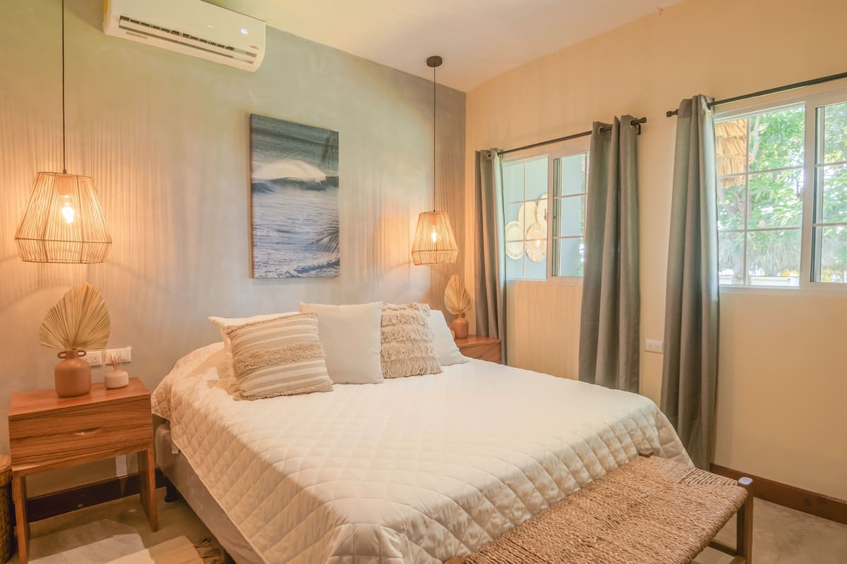 Un dormitorio con una cama grande, dos mesitas de noche, un aire acondicionado instalado en la pared, un cuadro y una ventana con cortinas.