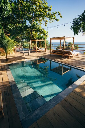 A wooden deck with a swimming pool, lounge chairs, and a view of the ocean. - El Xalli Hotel (Chiltiupan)